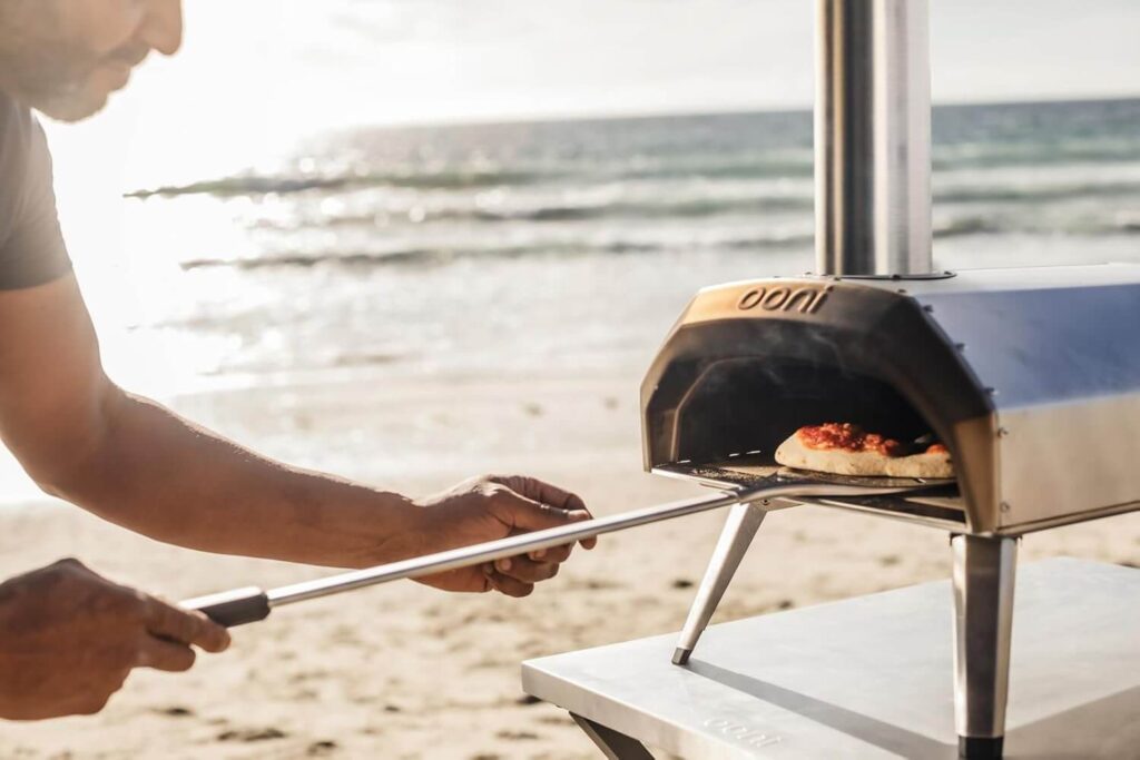 A man using a pizza peel to place a pizza into a portable Ooni Karu 12 pizza oven on a stand at the beach, with the ocean and sunset visible in the background
