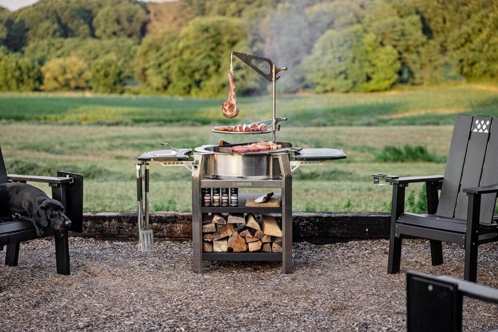 Breeo live fire grill set up outdoors with meat cooking over open flame, firewood storage, and patio seating