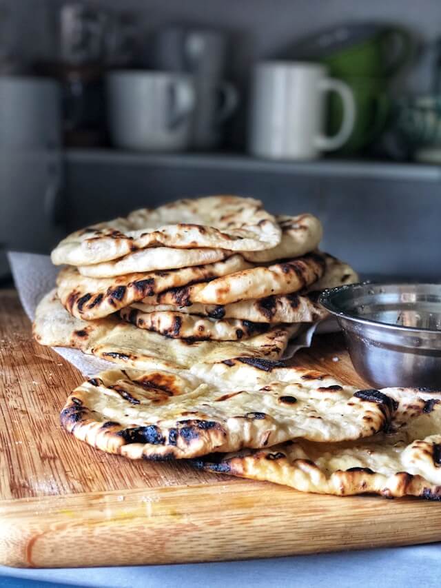 A close-up shot of a stack of freshly grilled flatbread on a wooden cutting board, with a small metal bowl next to it