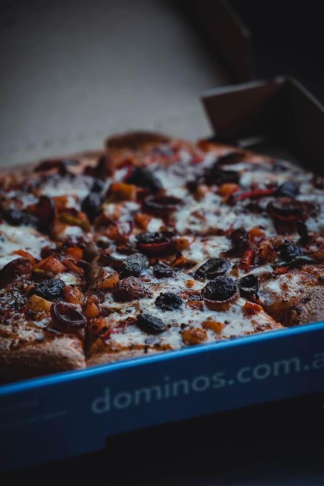 Fresh Domino’s large pizza sliced into eight cheesy pieces, topped with black olives, peppers, and melted mozzarella inside a blue delivery box