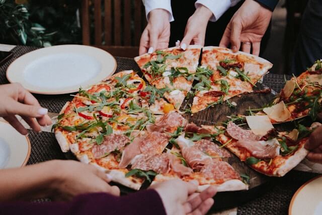 Friends sharing a large, gourmet wood-fired pizza topped with prosciutto, salami, arugula, and peppers at an outdoor restaurant table.