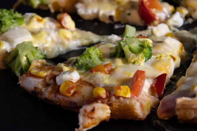Close-up of a rustic slice of homemade vegetarian pizza topped with melted mozzarella, fresh broccoli florets, sweet corn kernels, diced red peppers, and crumbled feta cheese on a dark background