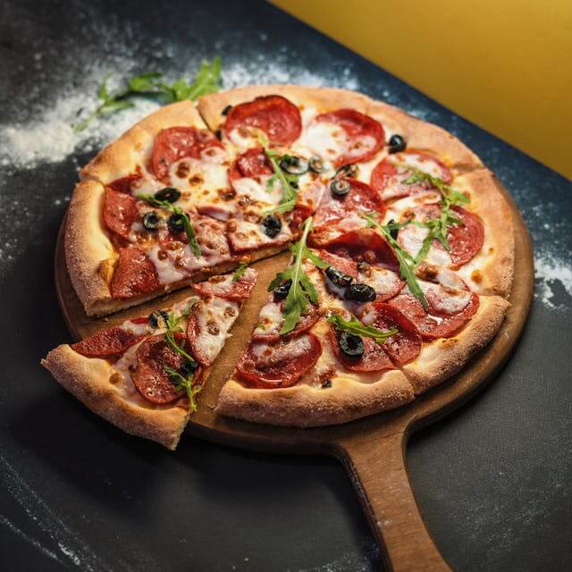 Freshly baked pepperoni pizza sliced into eight pieces, topped with black olives and arugula on a wooden board