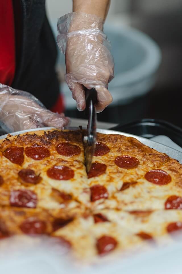 Close-up shot of a gloved hand using a pizza wheel to slice a freshly baked pepperoni and cheese pizza into quarters, emphasizing food preparation expertise in a commercial kitchen