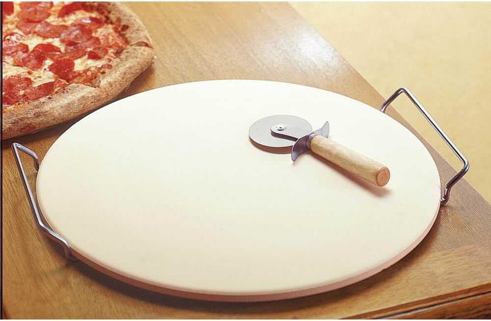 Ceramic pizza stone with metal handles and wooden-handled pizza cutter, next to a pepperoni pizza