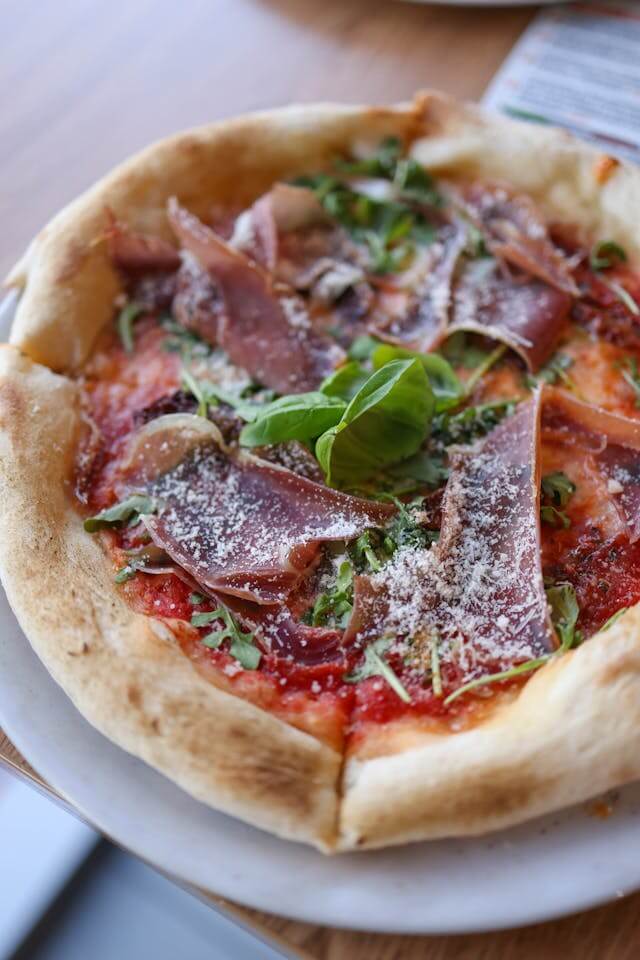 A freshly baked pizza topped with tomato sauce, prosciutto, arugula, and shaved parmesan on a white plate